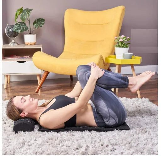 Woman practicing yoga on an acupressure yoga mat, sitting on a cozy rug with a yellow chair in the background.